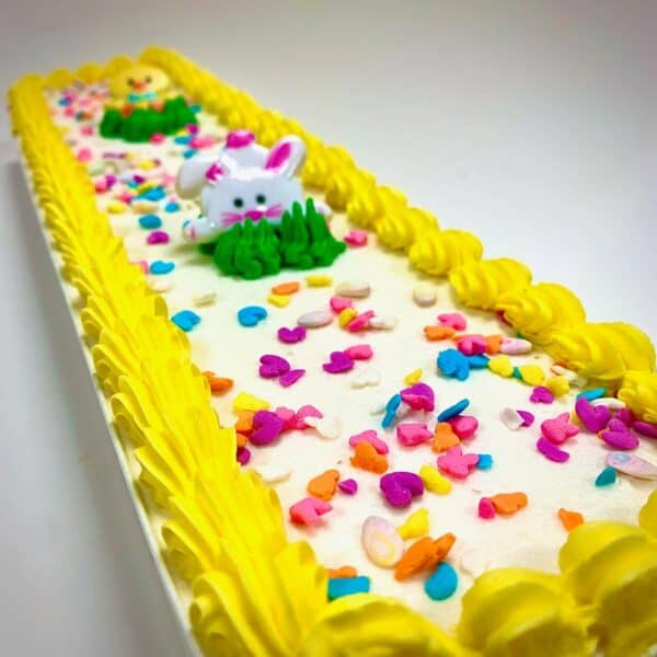 A yellow and white iced cake with a small decorative rabbit and rainbow sprinkles.